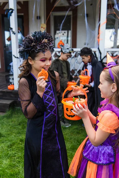 Smiling kid in halloween costume holding candy and bucket near friend in backyard — Stock Photo