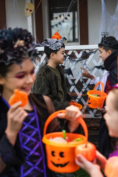 Asian boy in halloween costume holding bucket and pointing with finger at friend near blurred girls outdoors — Stock Photo