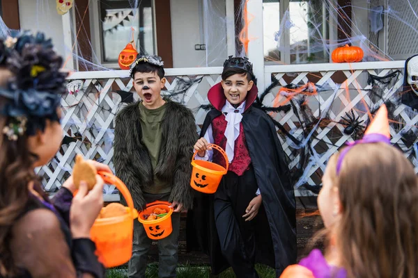 Asiático chicos mueca mientras sostiene halloween cubos cerca borrosa amigos al aire libre - foto de stock