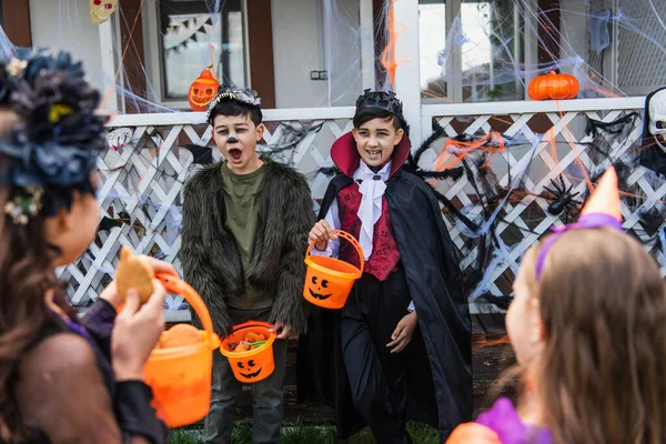 Asian boys grimacing while holding halloween buckets near blurred friends outdoors — Stock Photo