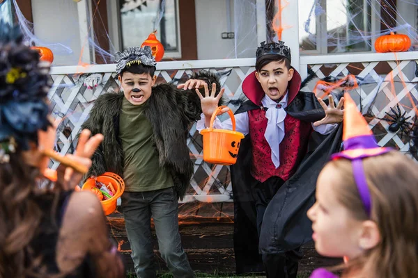 Enfants asiatiques en costumes d'Halloween tenant des seaux avec des bonbons près des amis flous dans la cour arrière — Photo de stock