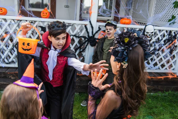 Preteen asiatique garçon tenant seau et effrayant amis pendant halloween fête dans arrière-cour — Photo de stock