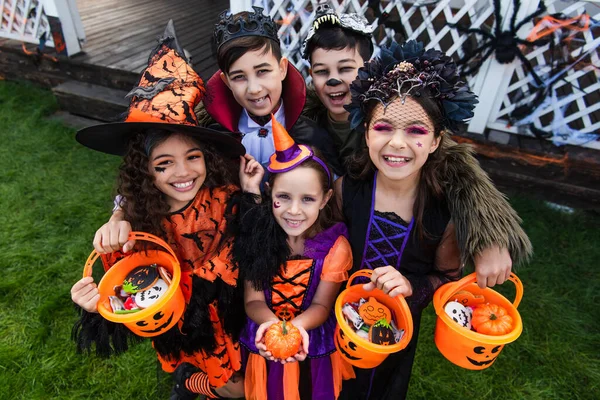 Vue grand angle des enfants souriants multiethniques tenant des seaux d'Halloween avec des bonbons et des citrouilles à l'extérieur — Photo de stock