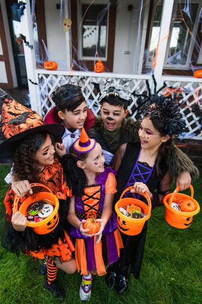 Cheerful multiethnic children in halloween costumes holding buckets with candies and pumpkin in backyard — Stock Photo