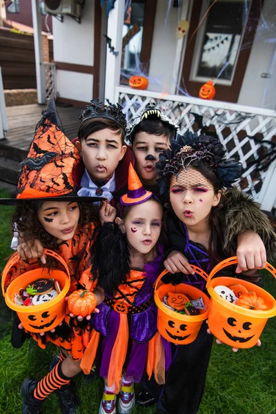 Multiethnic kids in costumes holding buckets with candies and grimacing at camera in backyard — Stock Photo