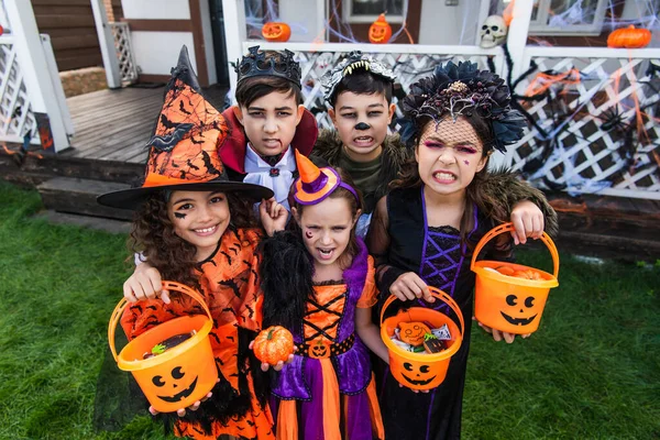 Multiethnic kids in halloween costumes holding buckets with candies and looking at camera in backyard — Stock Photo