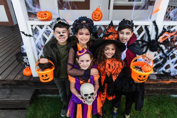 Blick aus der Vogelperspektive auf glückliche multiethnische Kinder in Halloween-Kostümen, die Eimer und Totenkopf in der Nähe des Hauses im Freien halten — Stockfoto