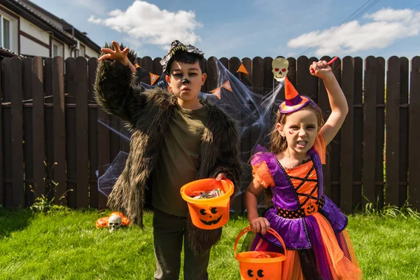 Мультиэтнические дети в костюмах на Хэллоуин держат ведра и гримасничают перед камерой на заднем дворе — стоковое фото