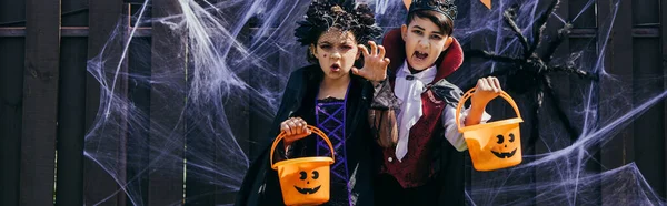 Multiethnic kids holding halloween buckets and grimacing at camera near decor on fence, banner — Stock Photo