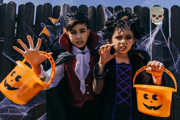 Interrazziale ragazzo e ragazza holding halloween secchi e smorfie a macchina fotografica vicino recinto in cortile — Foto stock