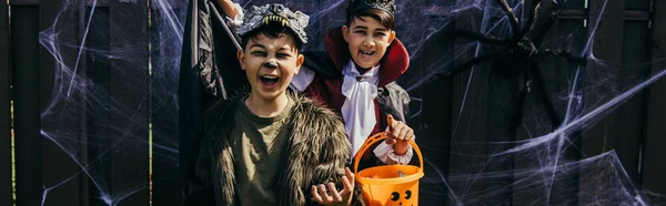 Crianças asiáticas em trajes de Halloween segurando balde com doces perto de teia de aranha na cerca no quintal, bandeira — Fotografia de Stock