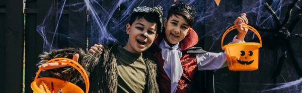 Positive asian kids in halloween costumes holding buckets and looking at camera outdoors, banner — Stock Photo