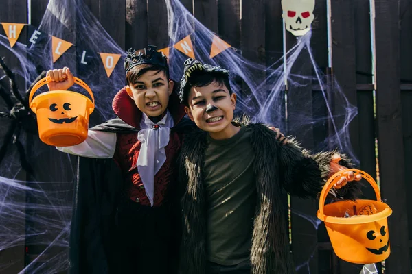 Asiatiques enfants avec maquillage grimacing à la caméra tout en tenant des seaux pendant Halloween fête en plein air — Photo de stock