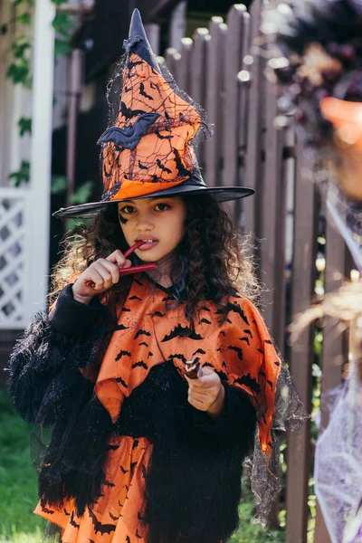 Preteen girl in witch hat eating candy during halloween celebration outdoors — Stock Photo