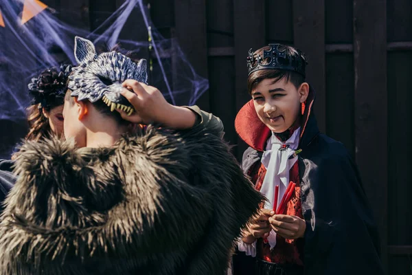 Улыбающийся азиатский мальчик в костюме Хэллоуина держит конфеты и смотрит в камеру рядом с друзьями на заднем дворе — стоковое фото