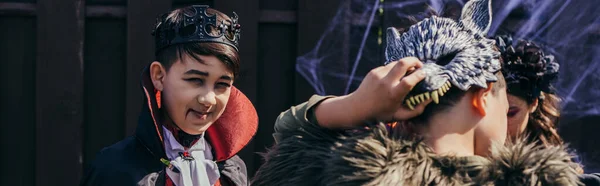 Sorridente bambino asiatico in costume e trucco guardando la fotocamera vicino agli amici durante la celebrazione di Halloween, banner — Foto stock