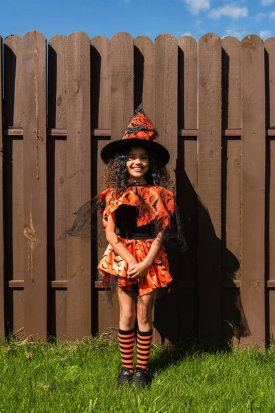 Chica sonriente en traje de Halloween mirando a la cámara cerca de valla en el patio trasero - foto de stock