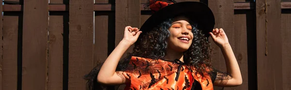 Cheerful girl in halloween costume holding witch hat near fence outdoors, banner — Stock Photo