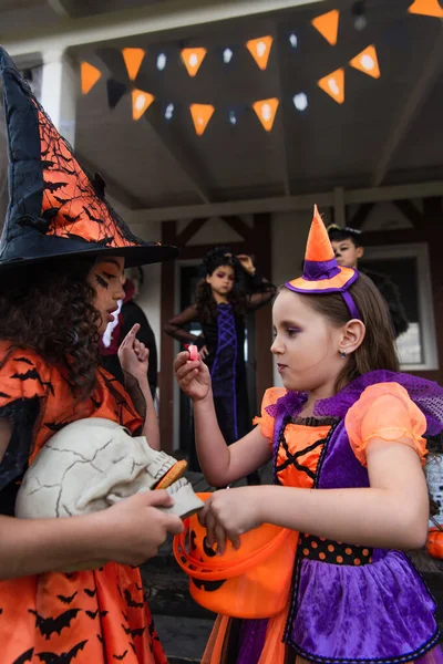 Chicas en disfraces de Halloween sosteniendo cráneo y truco o tratar cubo cerca borrosa interracial amigos - foto de stock