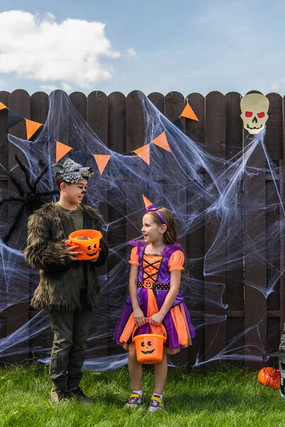 Улыбающиеся межрасовые дети в костюмах на Хэллоуин держат ведра с конфетками рядом с паутиной и декором на улице — стоковое фото