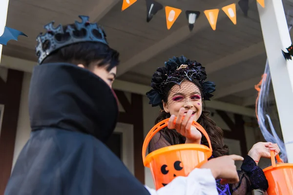 Lächelndes Mädchen hält Eimer neben asiatischem Freund im Halloween-Kostüm im Freien — Stockfoto