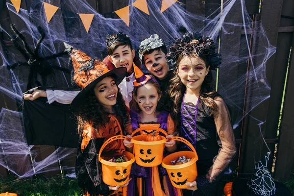 Positivo interracial crianças segurando baldes com doces perto halloween decoração no cerca ao ar livre — Fotografia de Stock