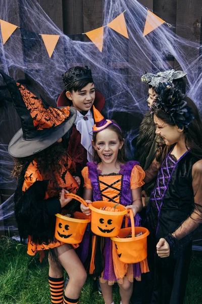 Chica alegre en traje sosteniendo cubo con caramelos cerca de amigos interracial y tela de araña en la cerca durante la fiesta de halloween — Stock Photo