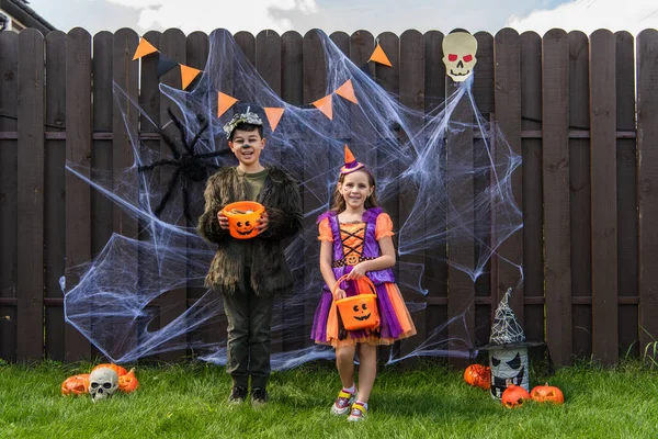 Sorrindo crianças multiétnicas segurando baldes com doces perto de decoração halloween na cerca no quintal — Fotografia de Stock