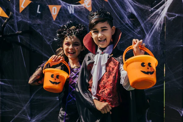 Smiling interracial preteen friends in costumes holding buckets during halloween party in backyard — Stock Photo