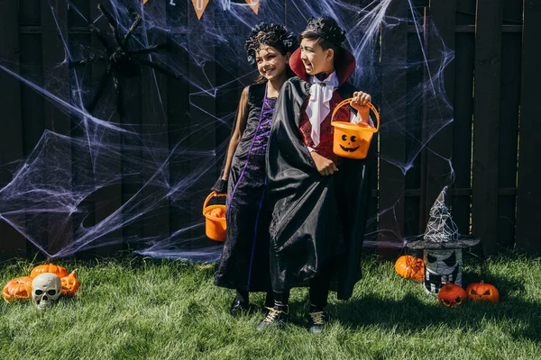 Lächelnde multiethnische Kinder in Kostümen mit Eimern in der Nähe von Halloween-Dekor am Zaun im Freien — Stockfoto