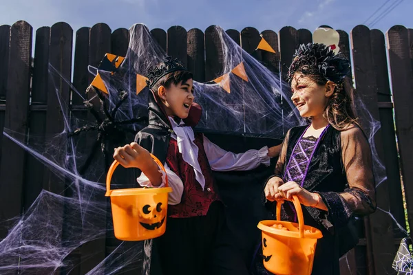 Asiático chico en halloween traje celebración cubo cerca sonriente amigo en patio trasero — Stock Photo