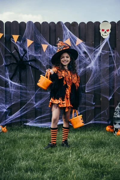 Усміхнена дівчина в капелюсі відьом тримає відра біля декору Хеллоуїна на паркані на задньому дворі — стокове фото