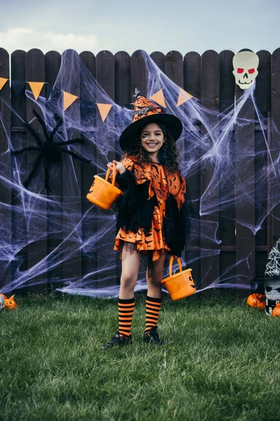 Ragazza sorridente in cappello da strega che tiene secchi vicino all'arredamento di Halloween sulla recinzione nel cortile — Foto stock