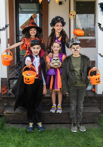 Весёлые многонациональные друзья в костюмах на Хэллоуин держат ведра с конфетами возле украшенного коттеджа — стоковое фото