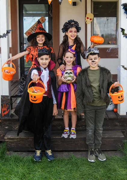 Cheerful multiethnic friends in halloween costumes holding buckets with candies near decorated cottage — Stock Photo