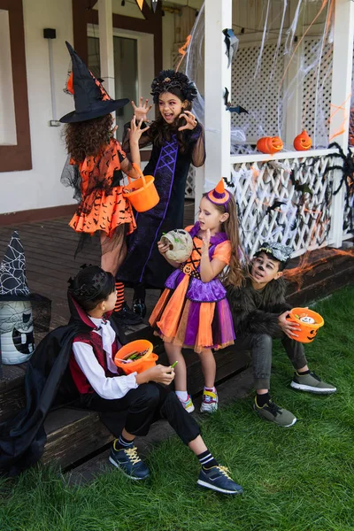 Asian boys sitting with halloween buckets near girls in witch costumes scaring each other — Stock Photo