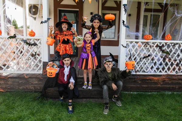 Мультиэтнические дети в костюмах на Хэллоуин гримасничают и показывают страшные жесты рядом с украшенным коттеджем — стоковое фото