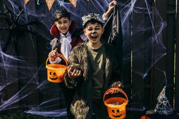 Asiático niños en trajes celebración de halloween cubos y muecas cerca de valla al aire libre - foto de stock