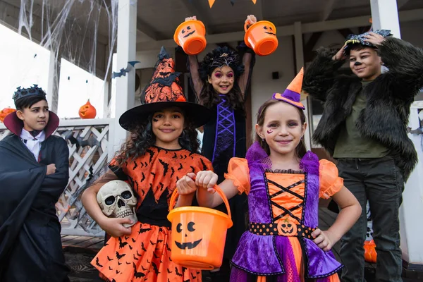 Alegre interracial niños en halloween trajes de pie con truco o tratar cubos cerca de casa - foto de stock