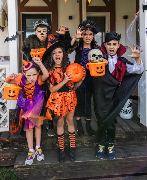 Niños multiétnicos en trajes de Halloween sosteniendo cubos y haciendo muecas en la cámara cerca de la casa - foto de stock
