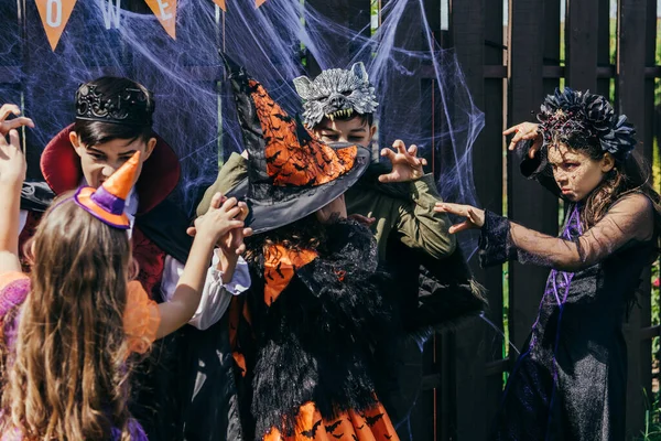 Niños multiétnicos en trajes festivos que se asustan mutuamente durante la fiesta de Halloween en el patio trasero - foto de stock