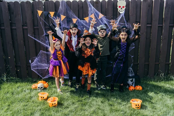 Interracial niños en halloween trajes de muecas en la cámara cerca de la decoración festiva en el patio trasero - foto de stock