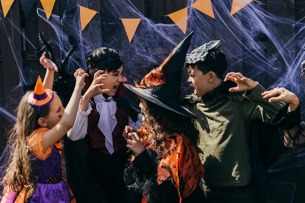 Interracial kids in costumes grimacing at each other near halloween decor in backyard — Stock Photo