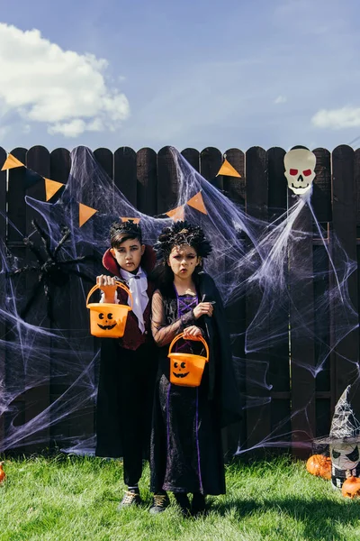 Мультиэтнические дети в костюмах на Хэллоуин держат ведра рядом с декором на заборе и траве на открытом воздухе — стоковое фото