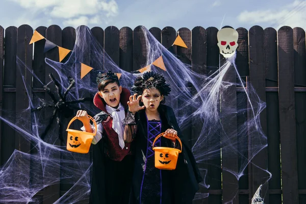 Niños interraciales con disfraces sosteniendo cubos de Halloween cerca de la tela de araña en la valla al aire libre - foto de stock