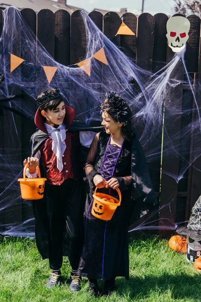 Asiático niño en halloween traje celebración cubo cerca amigo y decoración en patio trasero - foto de stock