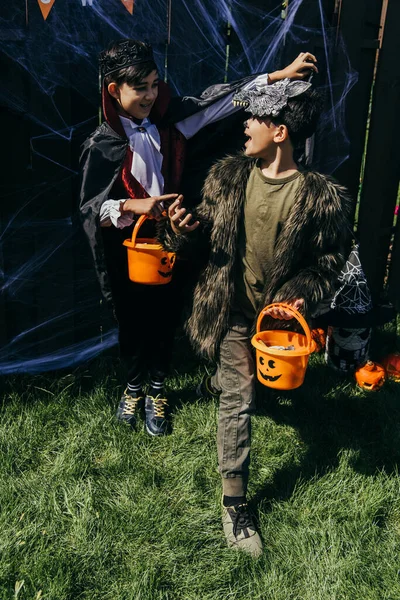 Cheerful asian boys in halloween costumes holding buckets with candies on grass in backyard — Stock Photo