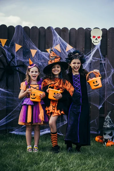 Smiling girls in costumes holding buckers with candies near halloween decor and spider web on fence outdoors — Stock Photo