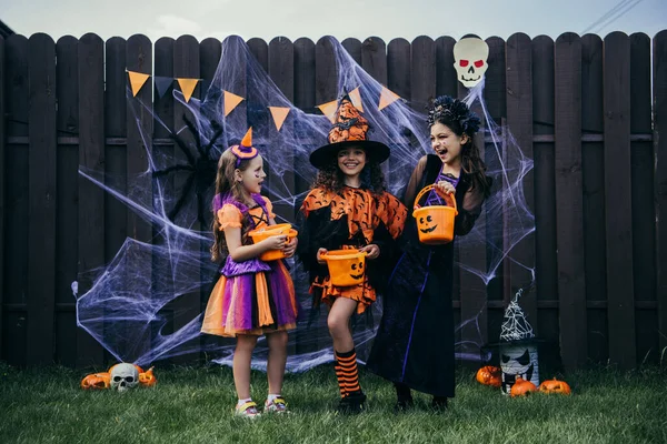 Cheerful preteen girls in costumes holding buckets near festive halloween decor on fence outdoors — Stock Photo