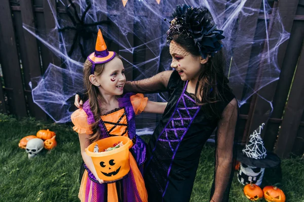 Lächelnde Mädchen mit Halloween-Make-up halten Bonbons im Eimer, während sie in der Nähe der Dekoration im Hinterhof stehen — Stockfoto